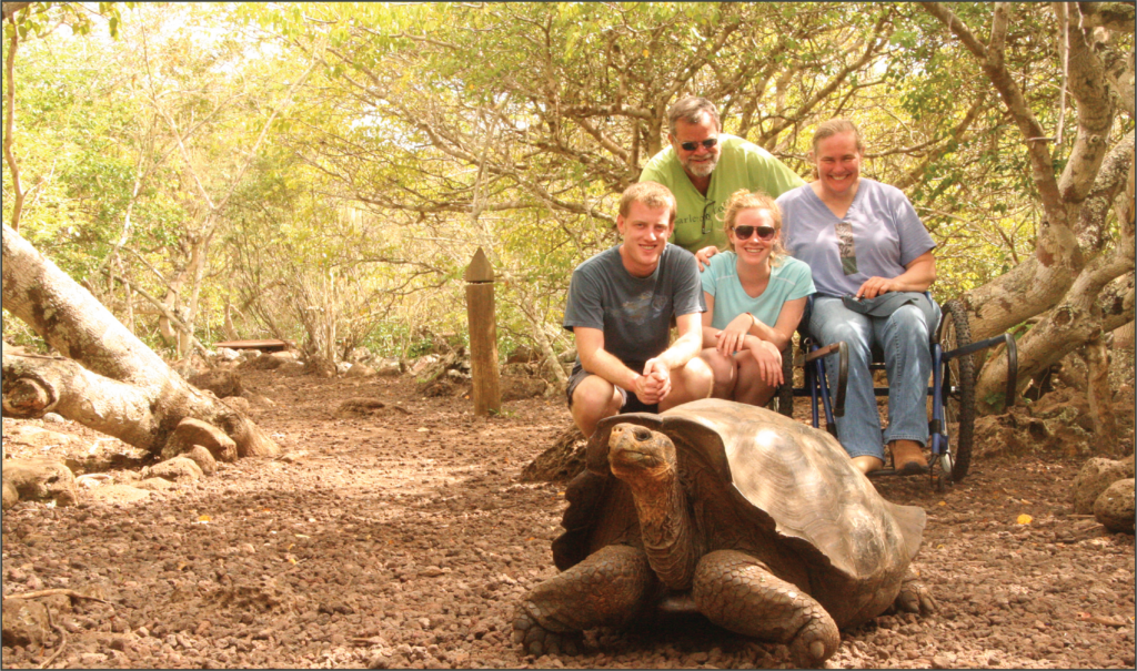 A family of four sits a few feet behind a tortoise, smiling. The mother is in a wheelchair, comfortably enjoying the tour and time with her love-ones.