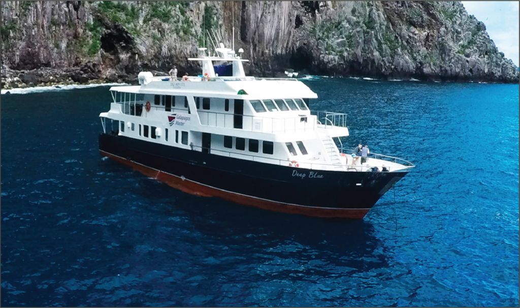 A bow and starboard view of a Galapagos ship near an island. The ship is navy, red and white.